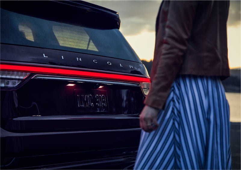 A person is shown near the rear of a 2023 Lincoln Aviator® SUV as the Lincoln Embrace illuminates the rear lights | Sheehy Lincoln of Richmond in Richmond VA